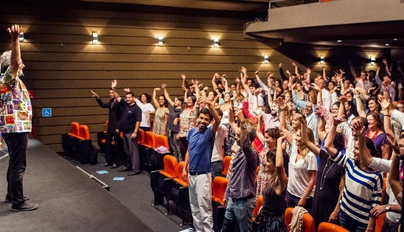 Imagem mostra plateia de um auditório, com pessoa de pé e com uma das mãos levantadas.