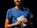 Foto mostra André Gravatá sorrindo com um microfone e segurando um papel nas mãos
