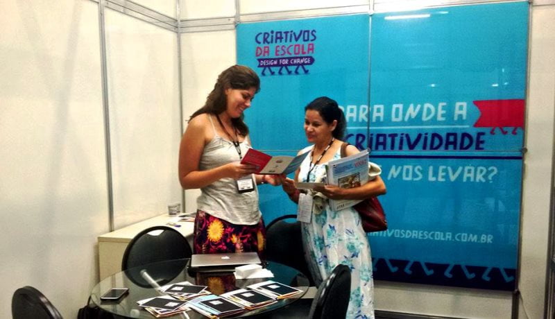 Mulheres em pé em frente a um banner do Criativos da Escola, conversando e vendo um livro.