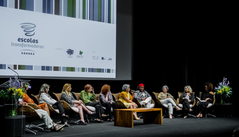 Pessoas sentadas em um palco. Atrás delas, aparece um telão multimídia.