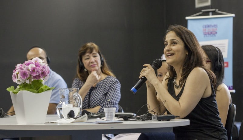 Mulheres sentadas em uma mesa. A da ponta direita segura um microfone, enquanto as outras prestam atenção.