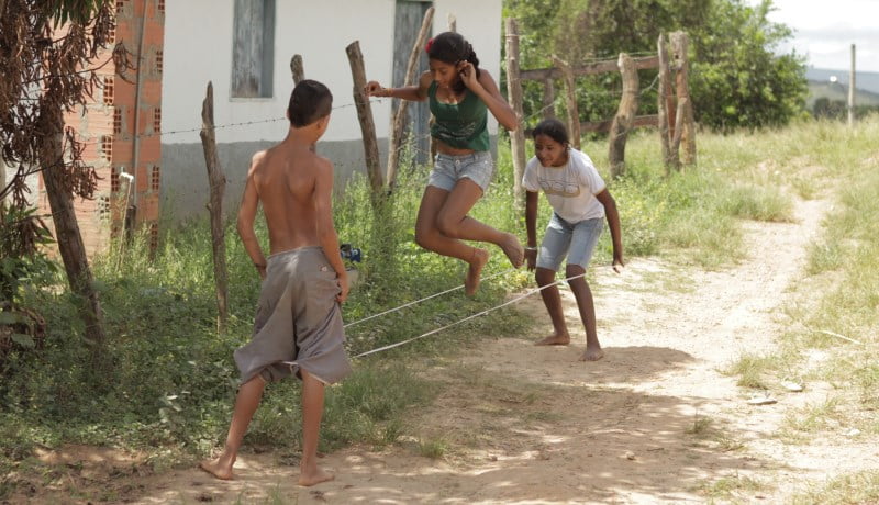 Crianças brincando de elástico em um caminho de terra e mato.