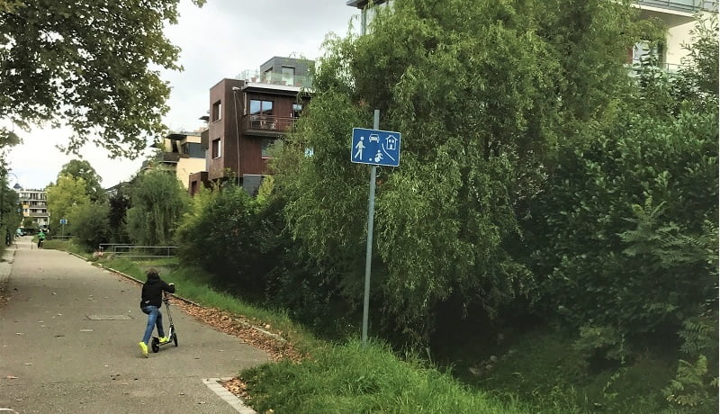 Foto de criança de costas andando de patinete pelas ruas do Bairro de Vauban, na Alemanha, ao lado do asfalto, muita natureza