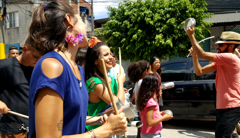 Foto mostra pessoas se divertindo durante desfile de carnaval. Em primeiro plano, duas mulheres segurando baquetas