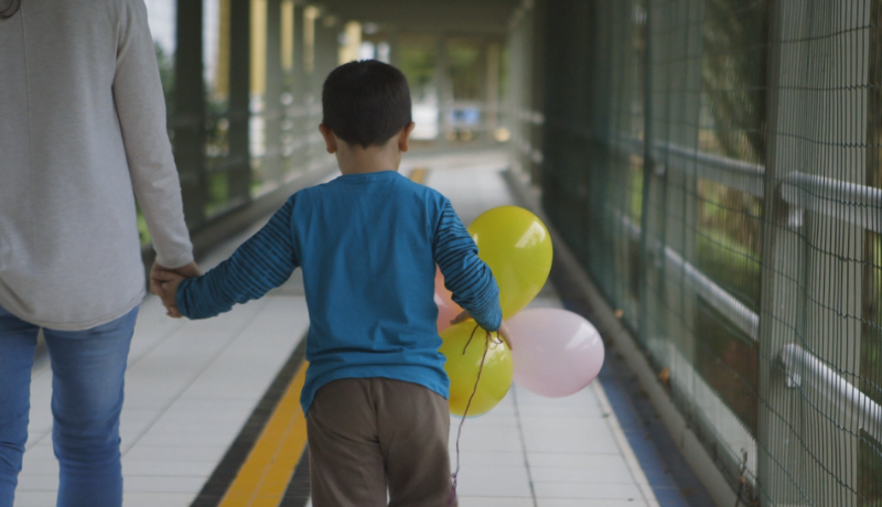 Imagem de um adulto e um menino com bexigas amarelas e rosas na mão. Os dois estão de costas andando por um corredor.