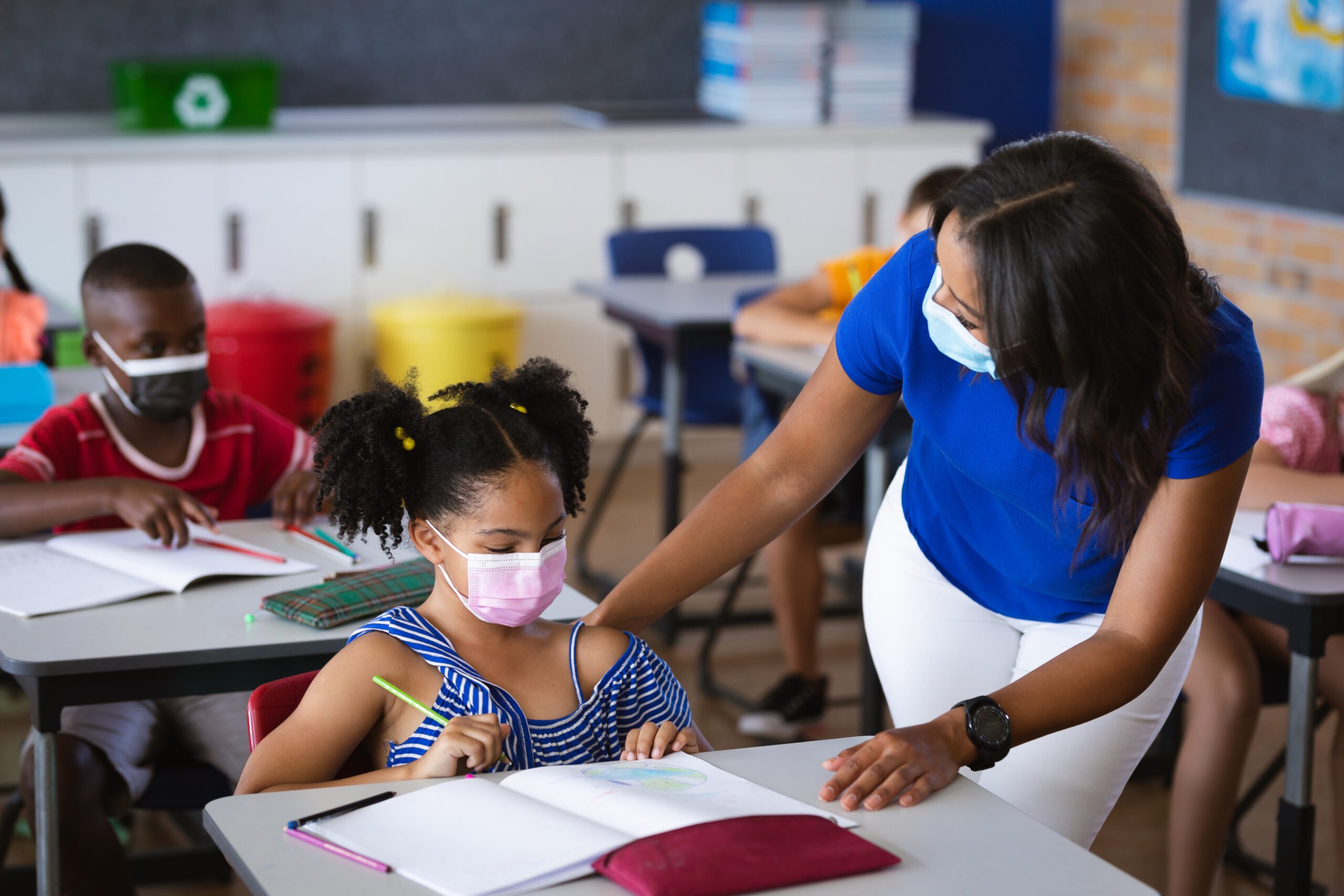 Foto na volta às aulas. Uma professora auxilia uma criança que está sentada em uma carteira escolar fazendo a lição. Todos usam máscaras de proteção.