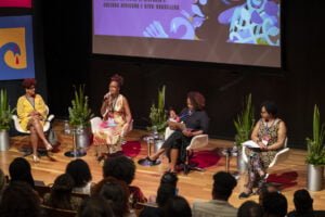 Nilma Lino Gomes, Márcia Lima, Lucimar Dias e Barbara Carine estão sentadas em um palco, debatendo de frente para uma plateia