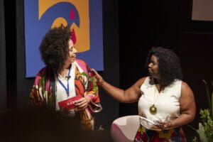 Beatriz Benedito e Tânia Portella olham uma para outra, de pé em um palco